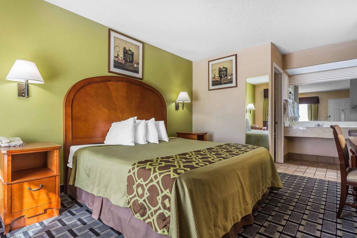 A hotel room with a large bed, two bedside tables, a nightstand, a bathroom visible through an open door, and a chair.