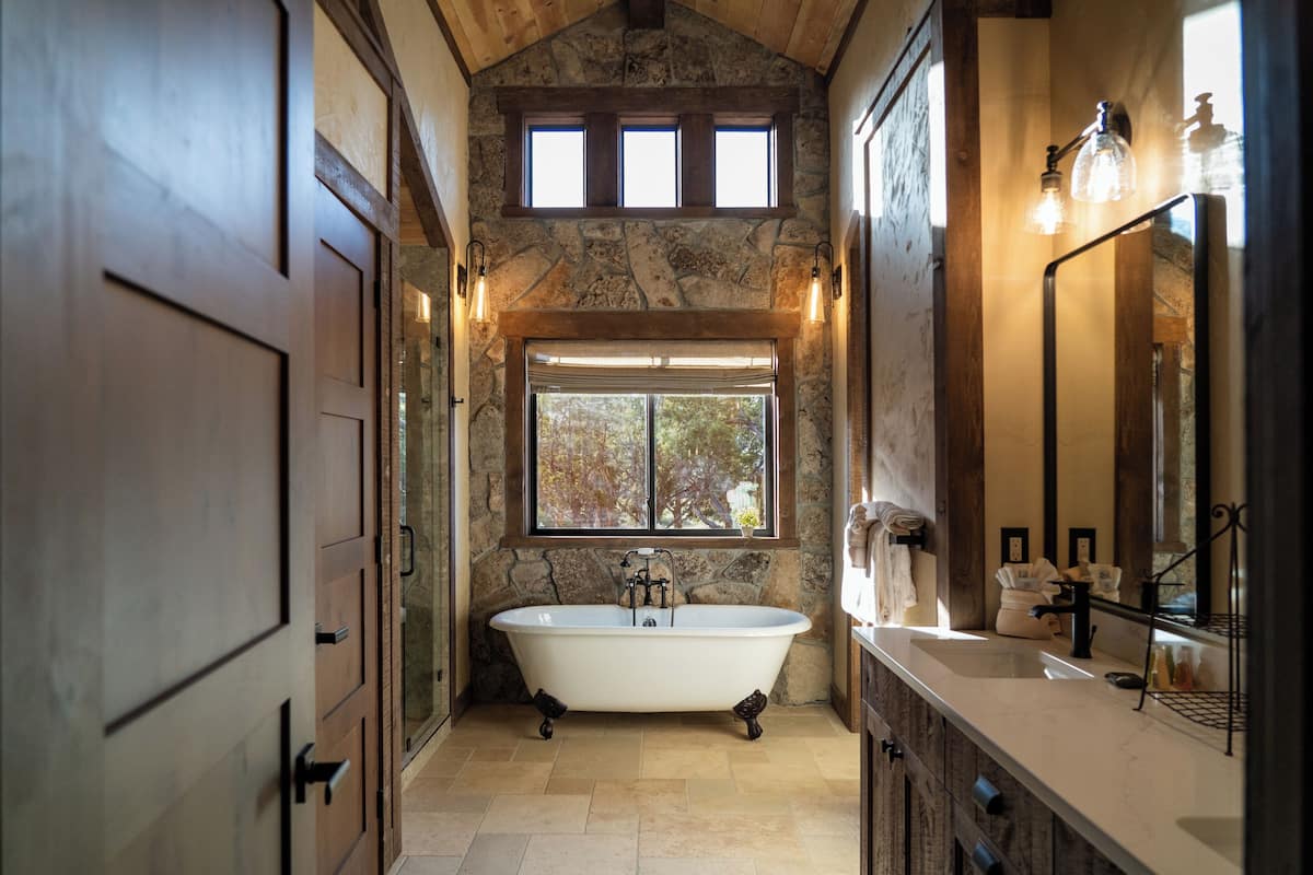 A bathroom with a freestanding bathtub, a large window, stone walls, and a wooden ceiling.