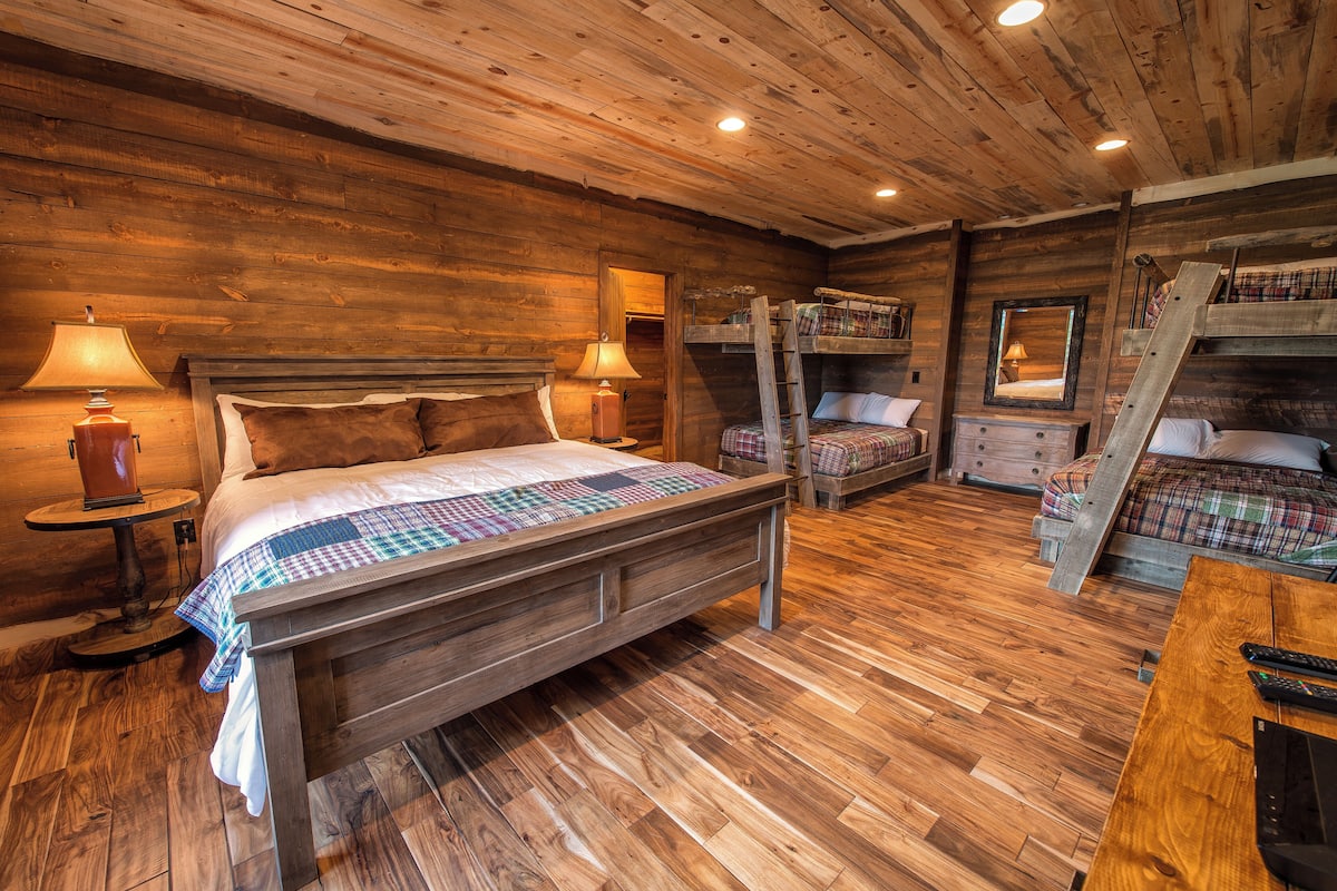 A rustic wooden room with a bed, bunk beds, a table, and lamps.