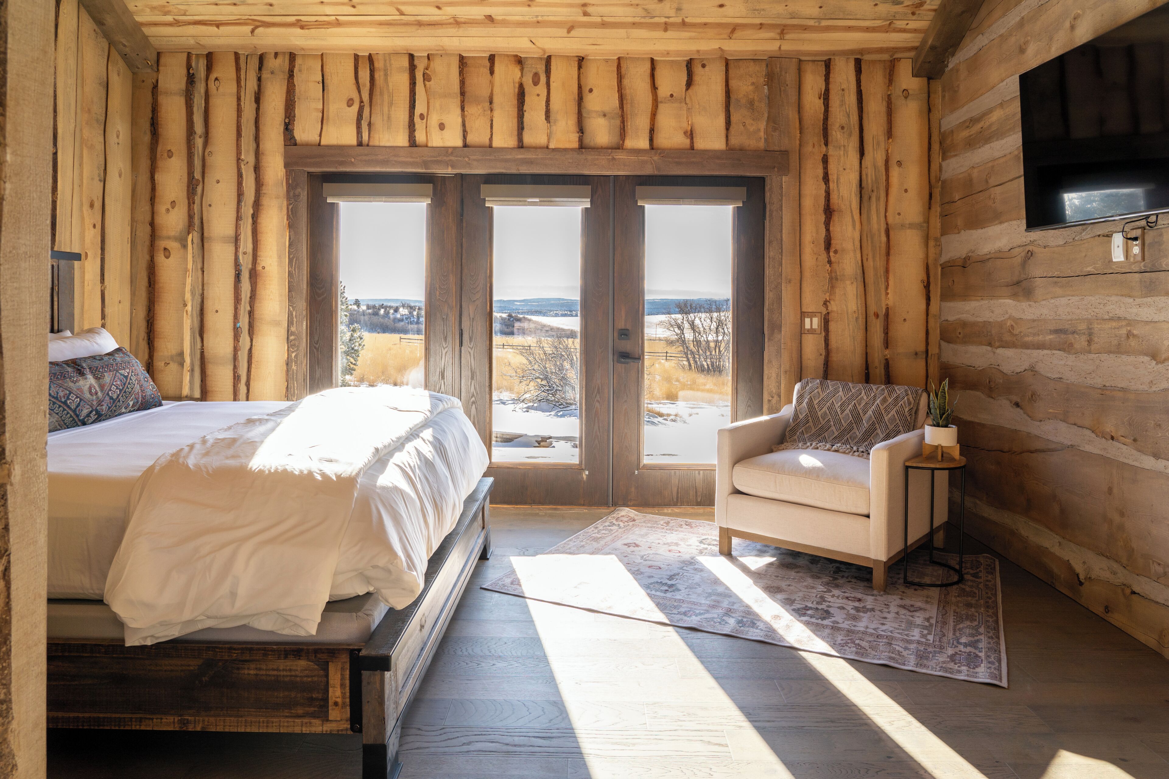 Een slaapkamer in een blokhut met een bed, een stoel, een televisie en uitzicht op de natuur.