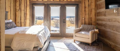 A log cabin bedroom with a bed, a chair, a TV, and a view of the outdoors.
