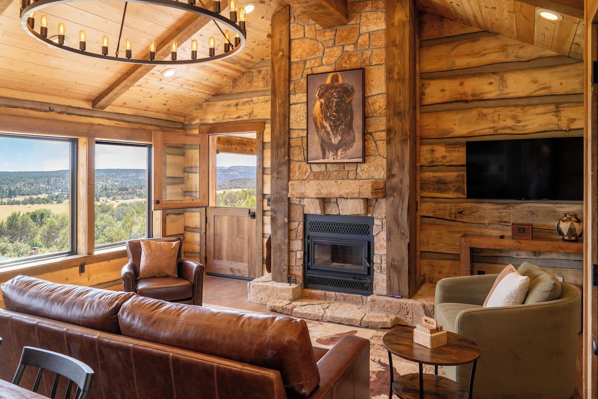 A log cabin interior with a stone fireplace, leather furniture, a television, and large windows offering a view of the outdoors.