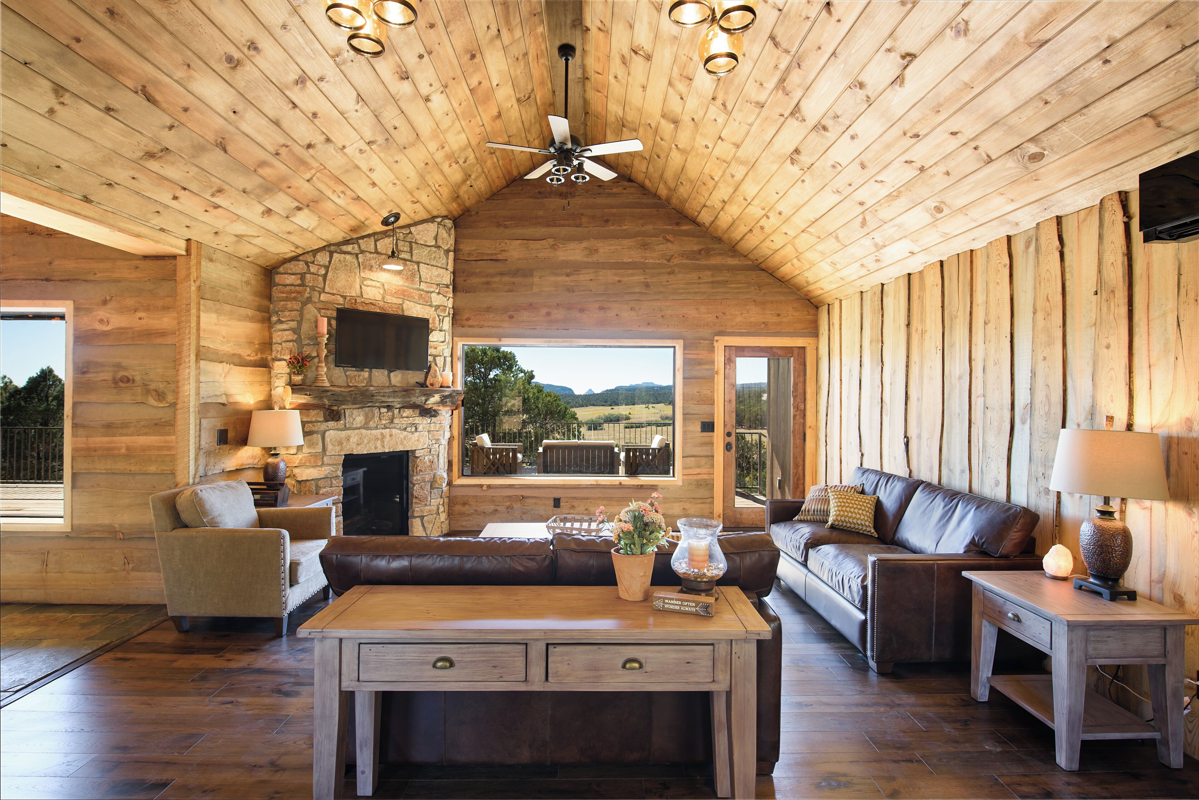A log cabin interior with a stone fireplace, leather sofas, a wooden coffee table, and a ceiling fan.