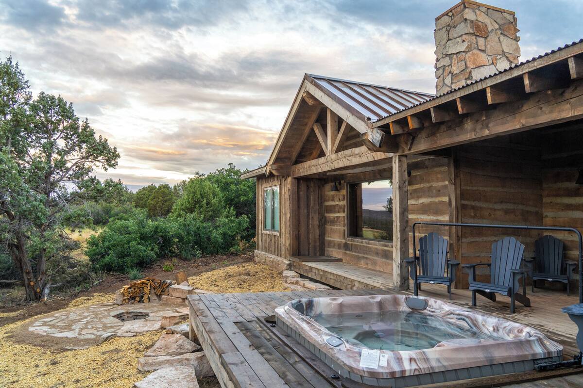 A wooden cabin with a hot tub, Adirondack chairs, and a stone chimney.