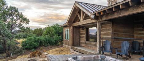 A wooden cabin with a hot tub, Adirondack chairs, and a stone chimney.