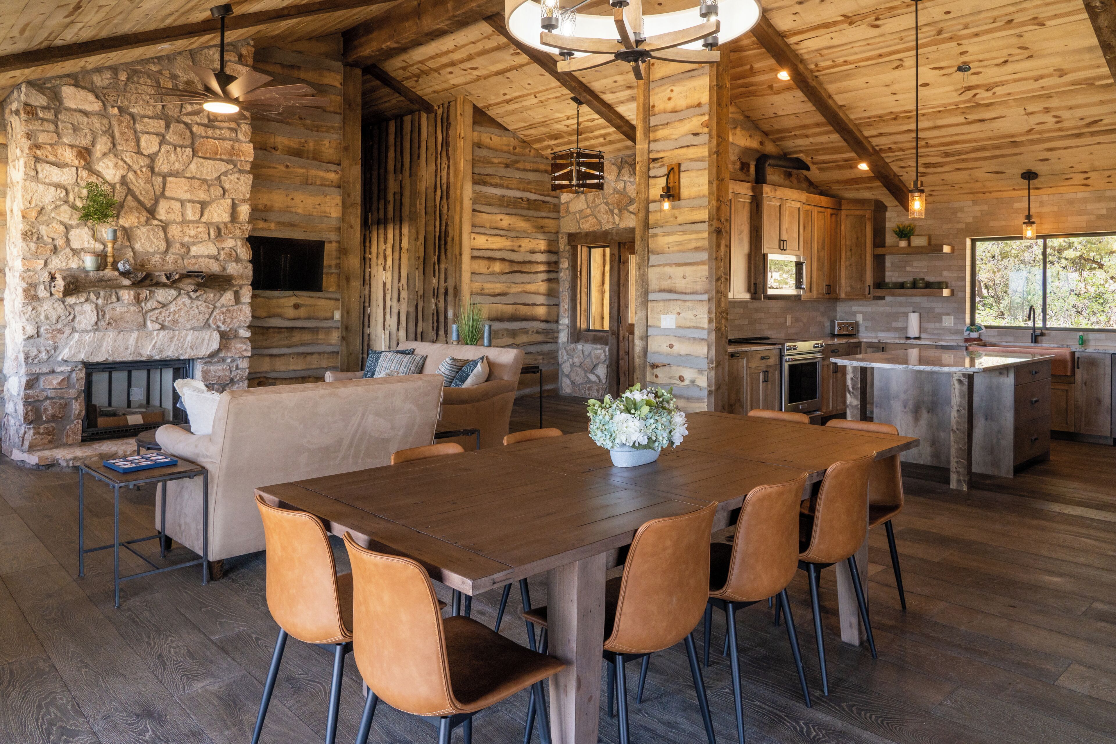 A rustic cabin interior with a stone fireplace, wooden dining table, and a kitchen area.