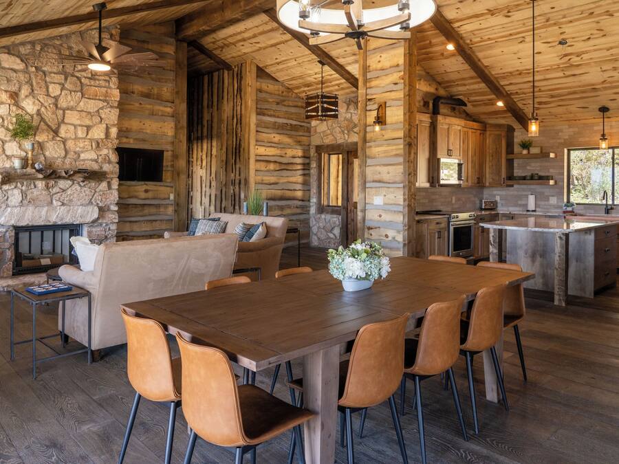 A rustic cabin interior with a stone fireplace, wooden dining table, and a kitchen area.