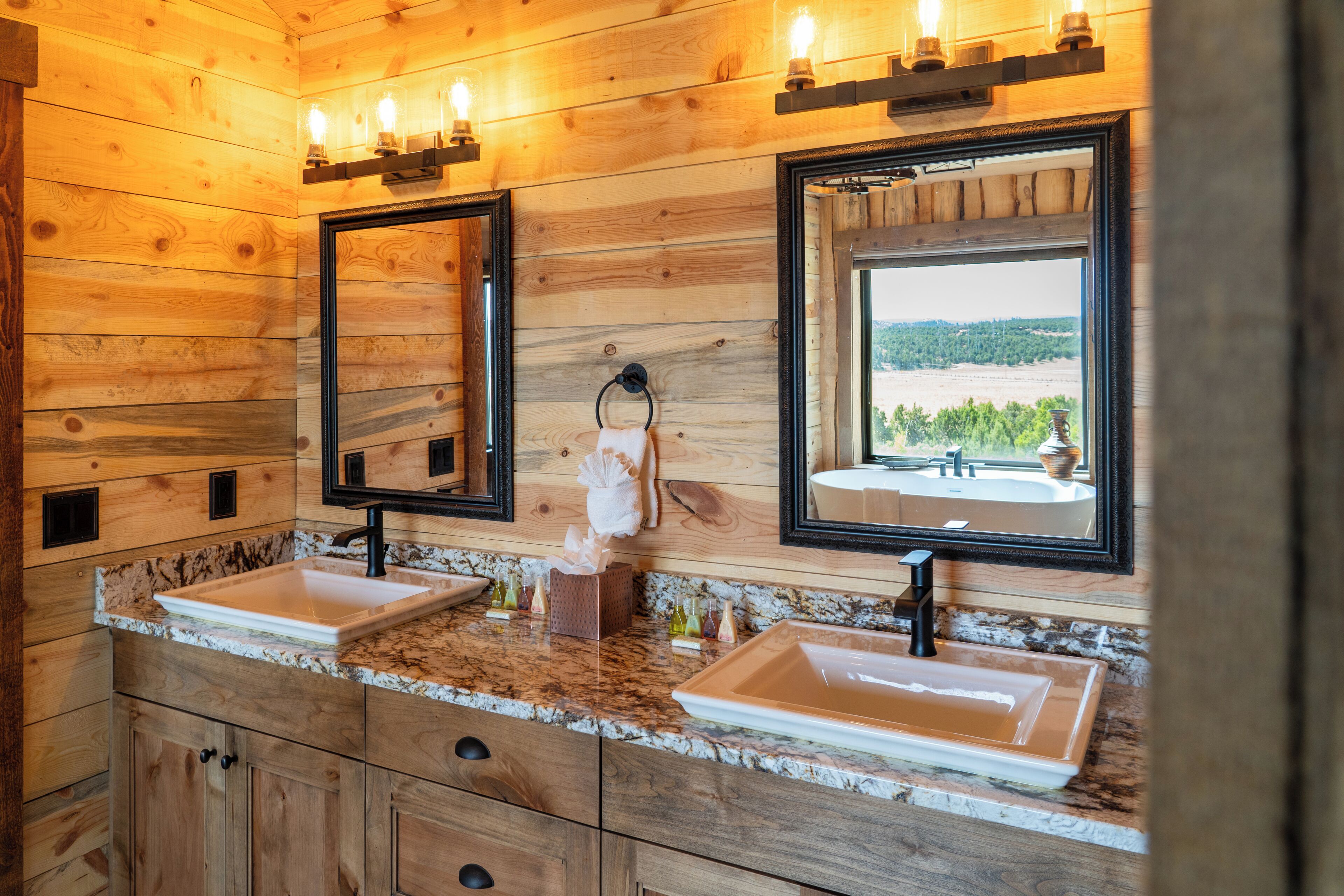 Une salle de bain rustique, avec deux lavabos, des miroirs et une fenêtre donnant sur un paysage agréable.