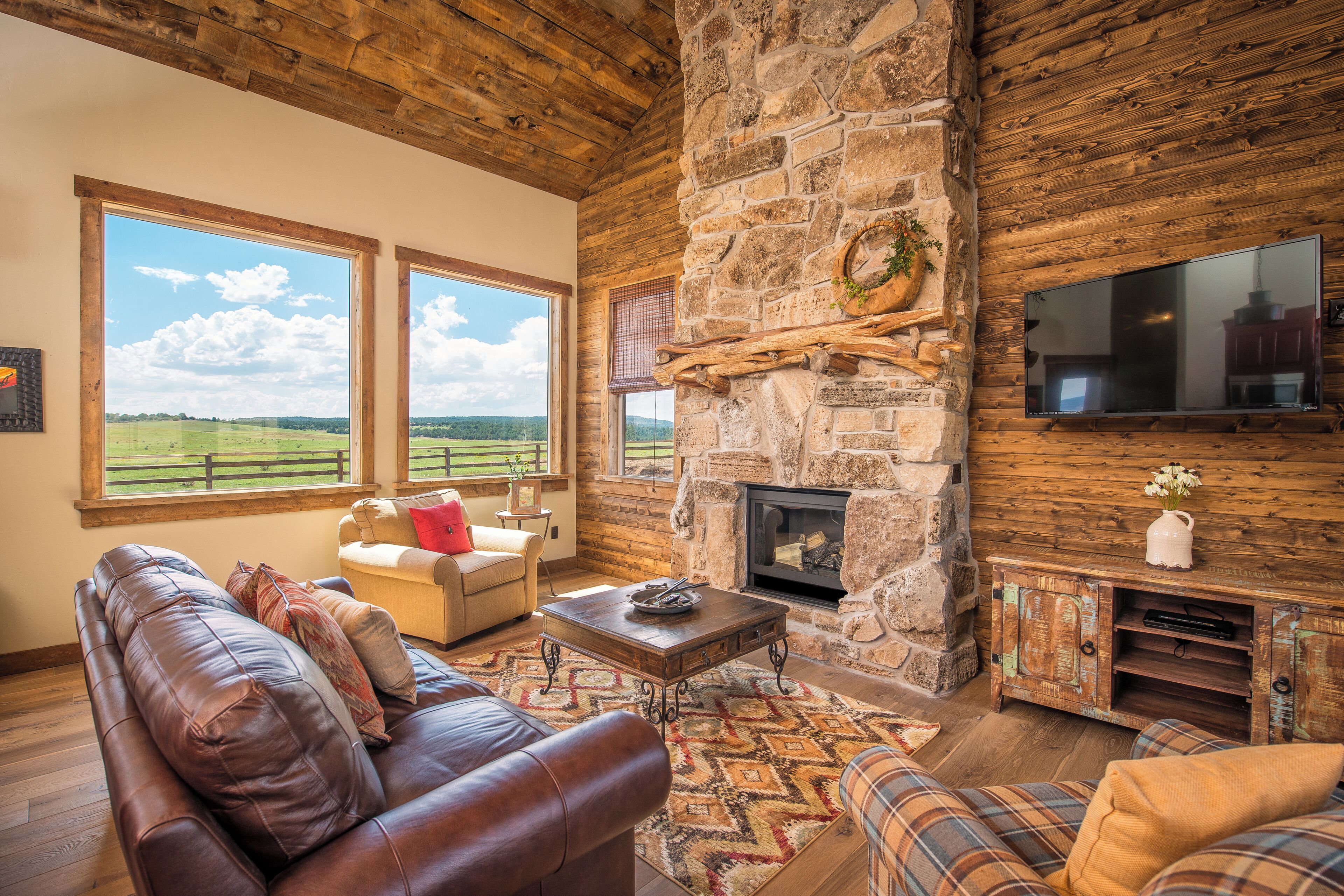 A cozy living room with a stone fireplace, leather sofa, and large windows overlooking a field.