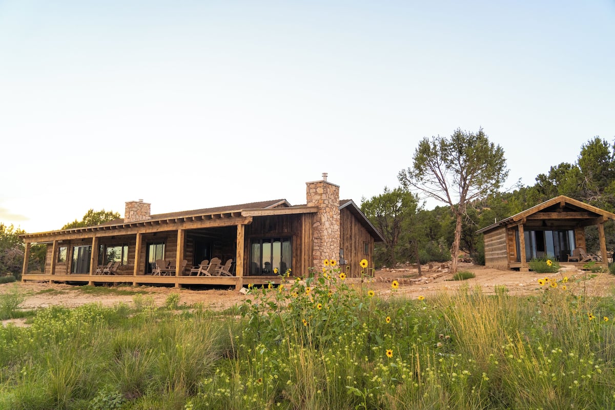 A rustic cabin with a stone chimney, a covered porch with chairs, and a small wooden structure.