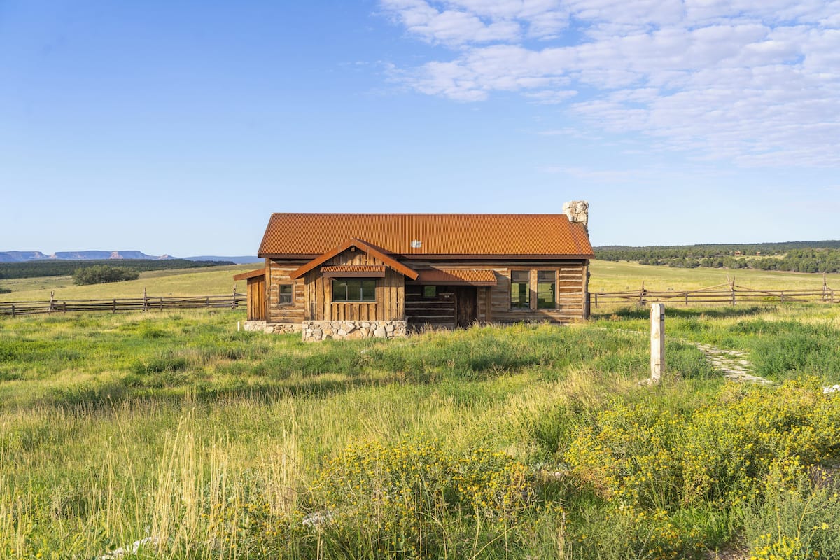 A rustic log cabin with a metal roof, surrounded by a grassy field and a wooden fence.