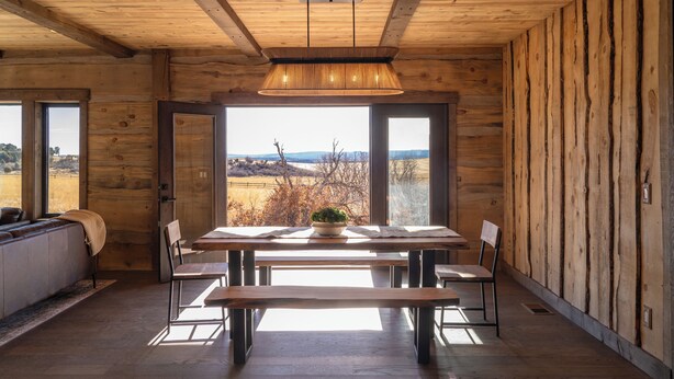 Un interior de madera con una mesa de comedor, sillas, un banco y vistas a través de amplios ventanales.