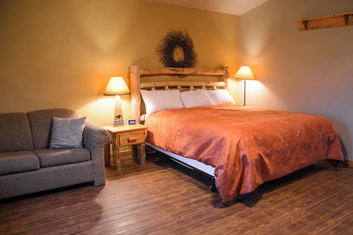 A bedroom with a bed, a wooden headboard, two bedside lamps, a nightstand, and a gray sofa.