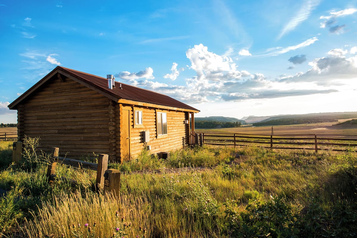 A wooden cabin with a metal roof, surrounded by tall grass and a wooden fence.