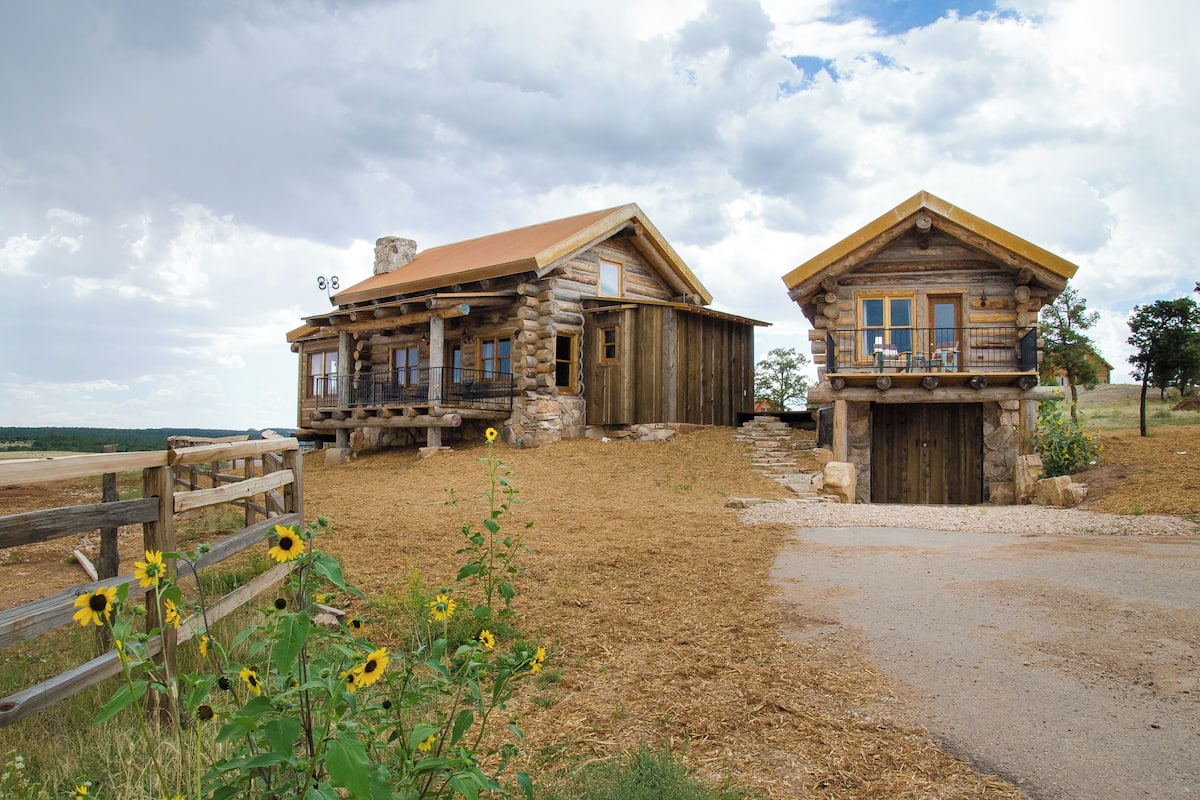 A rustic log cabin with a stone foundation, a wooden porch, and a clear sky.