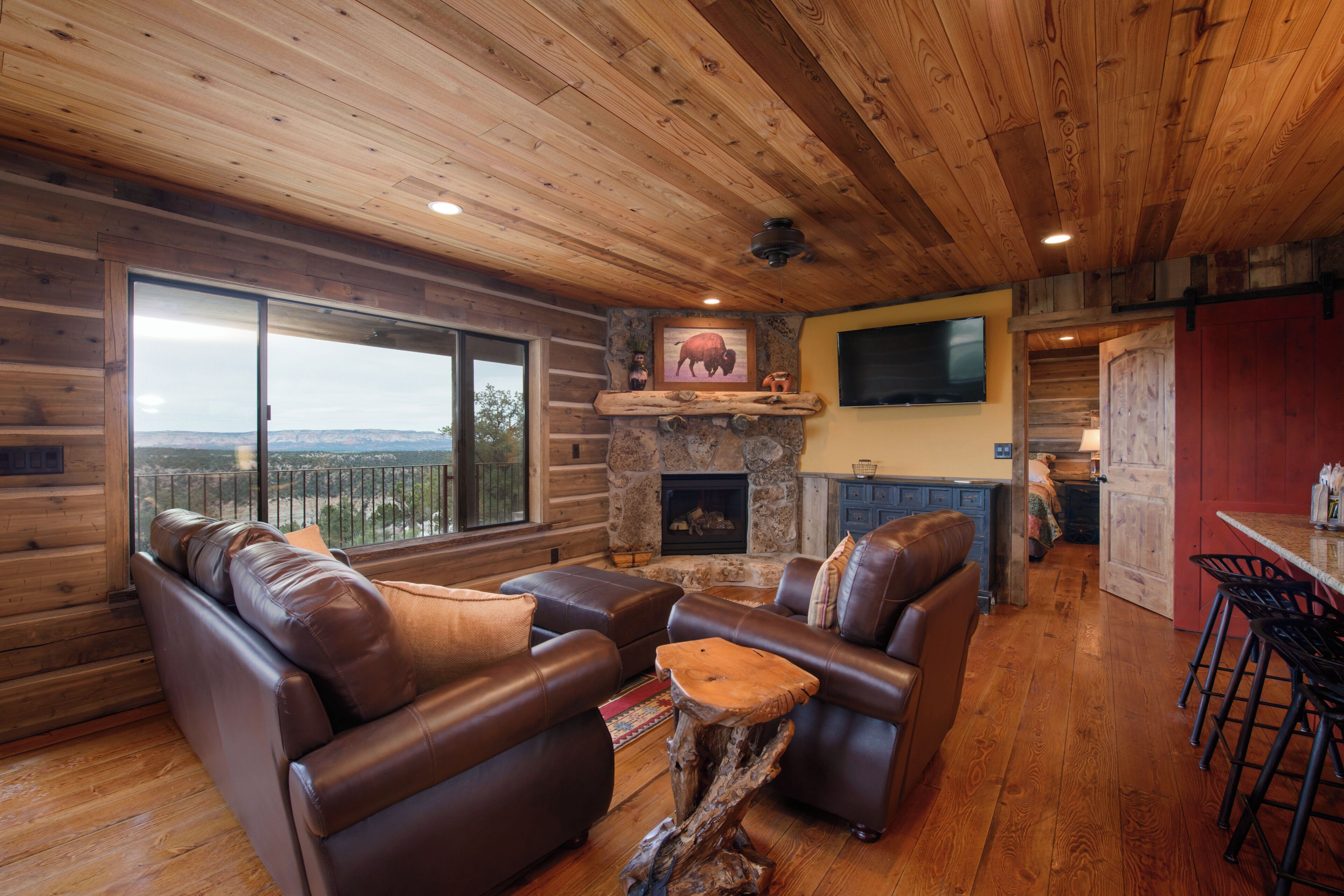 A log cabin interior with a stone fireplace, leather sofas, a bar area, and a large window offering a view.