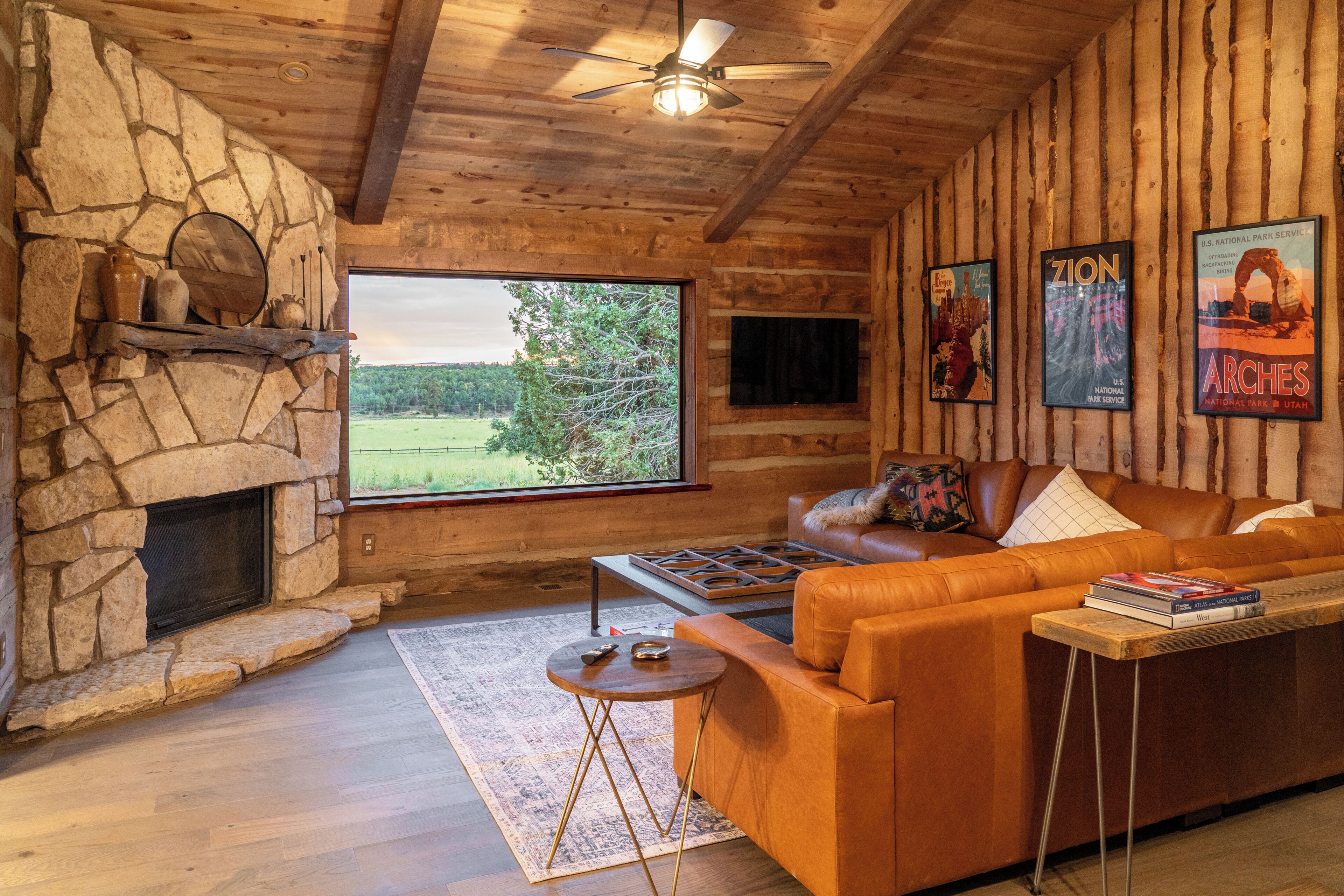 A log cabin interior with a stone fireplace, a leather sofa, a coffee table, and framed posters on the wall.