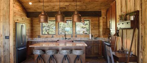 Une cuisine rustique aménagée dans une cabane en bois, avec un îlot central, des tabourets de bar, un réfrigérateur et des luminaires suspendus.