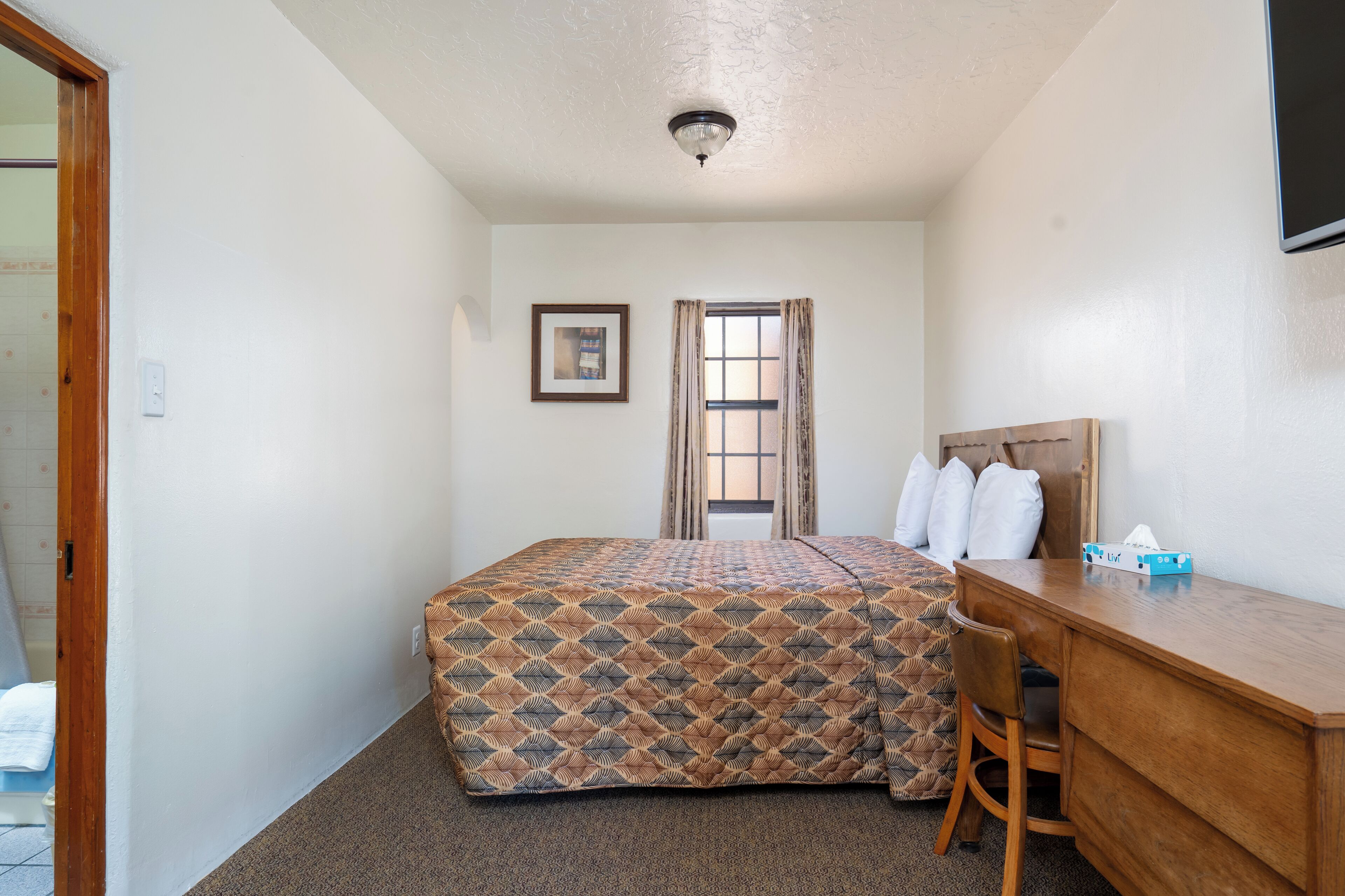A hotel room with a bed, a desk, a chair, a window with curtains, a framed picture on the wall, and a ceiling light.