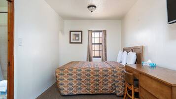A hotel room with a bed, a desk, a chair, a window with curtains, a framed picture on the wall, and a ceiling light.