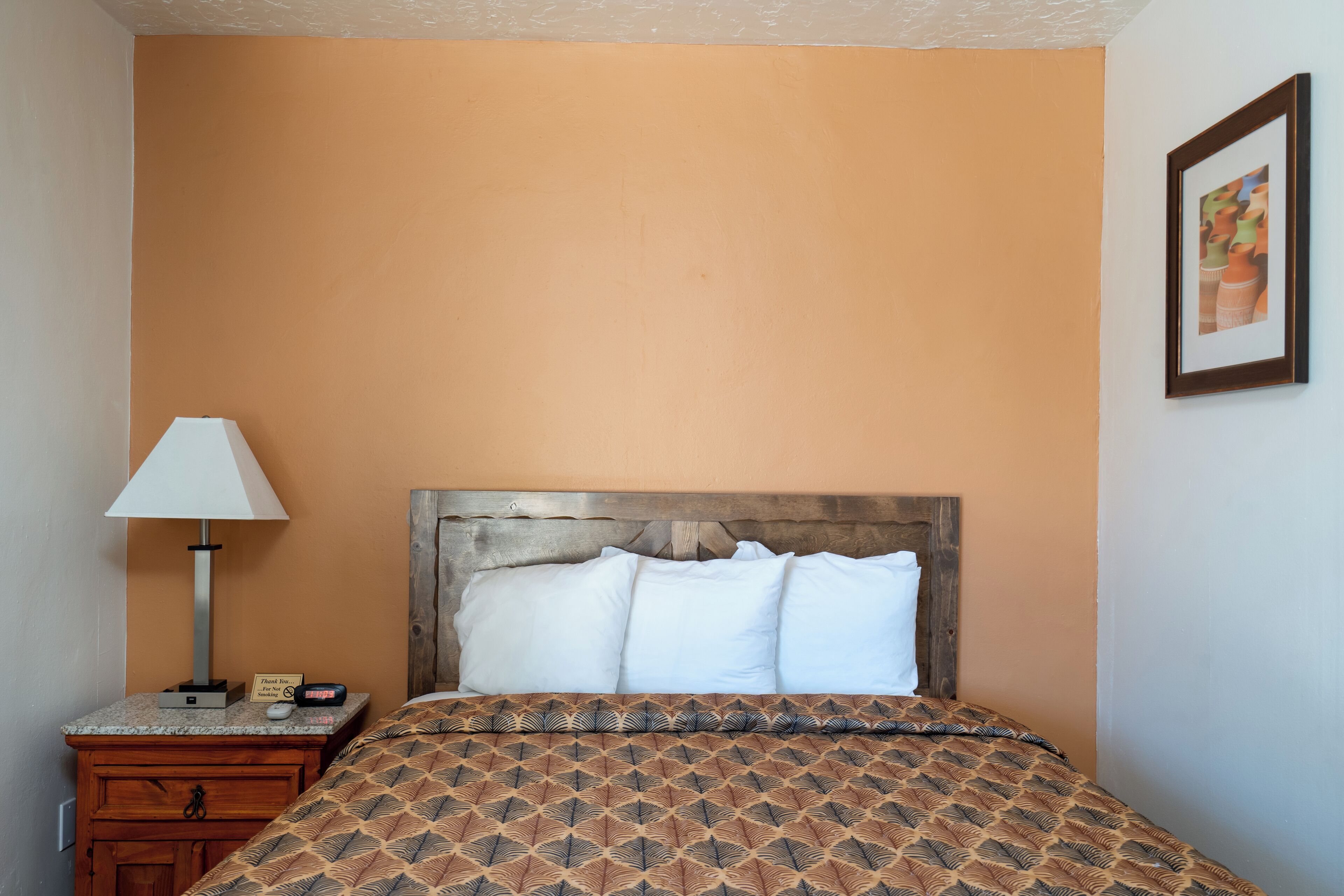 Uma cama bem arrumada com colcha estampada, cabeceira de madeira e mesa de cabeceira com abajur e uma pequena placa.