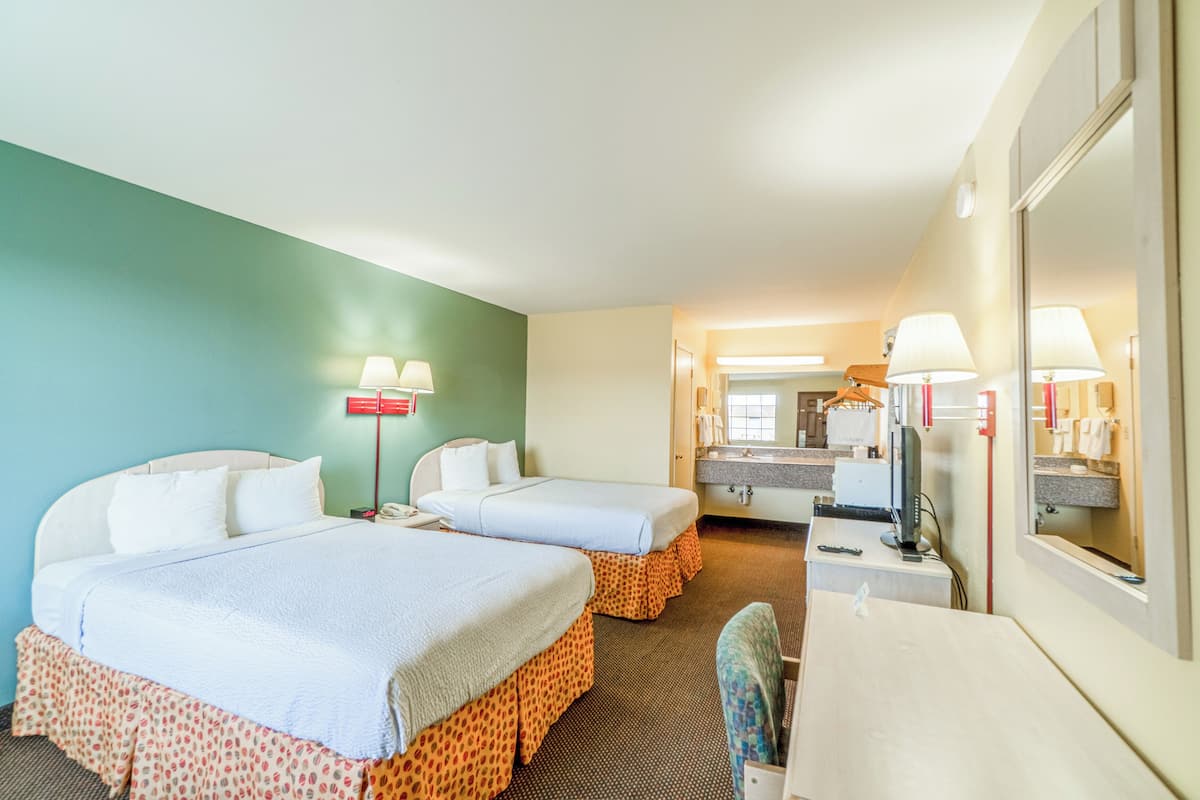 A hotel room with two beds, a desk, a mirror, and a bathroom visible in the background.