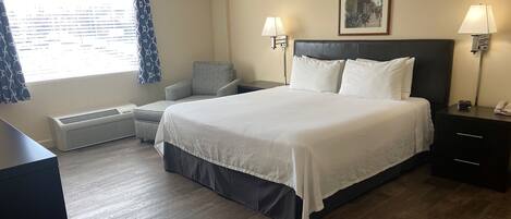 Ein Hotelzimmer mit einem großen Bett, zwei Nachttischlampen, einem Sessel, einem Schreibtisch, einem Heizkörper und einem fenster mit Vorhängen.
