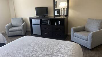 A hotel room with a bed, two armchairs, a dark wooden dresser with a television and microwave, and a large mirror.