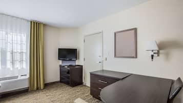 Quarto de hotel com cama, escrivaninha, cadeira, televisão, guarda-roupa, luminária e janela com cortinas.