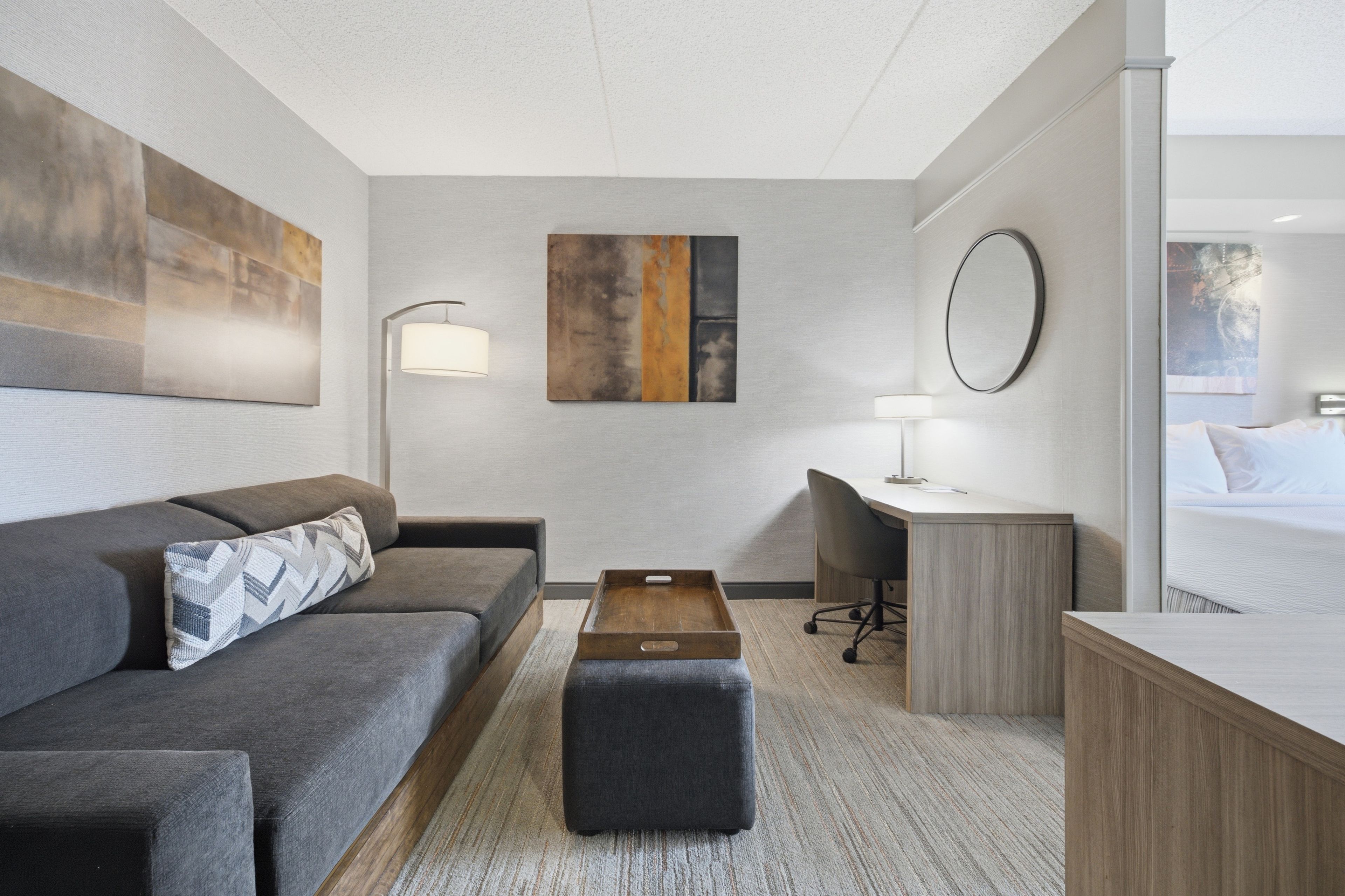 Une chambre d’hôtel moderne avec un canapé gris, une table d’appoint en bois, un bureau avec une chaise, un grand miroir et un lit.