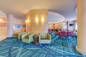 Un vestíbulo de hotel con diseño moderno, que presenta un techo curvo, un pilar central grande y suelo alfombrado. Hay varias zonas de estar con sillas y mesas, y un televisor montado en la pared.