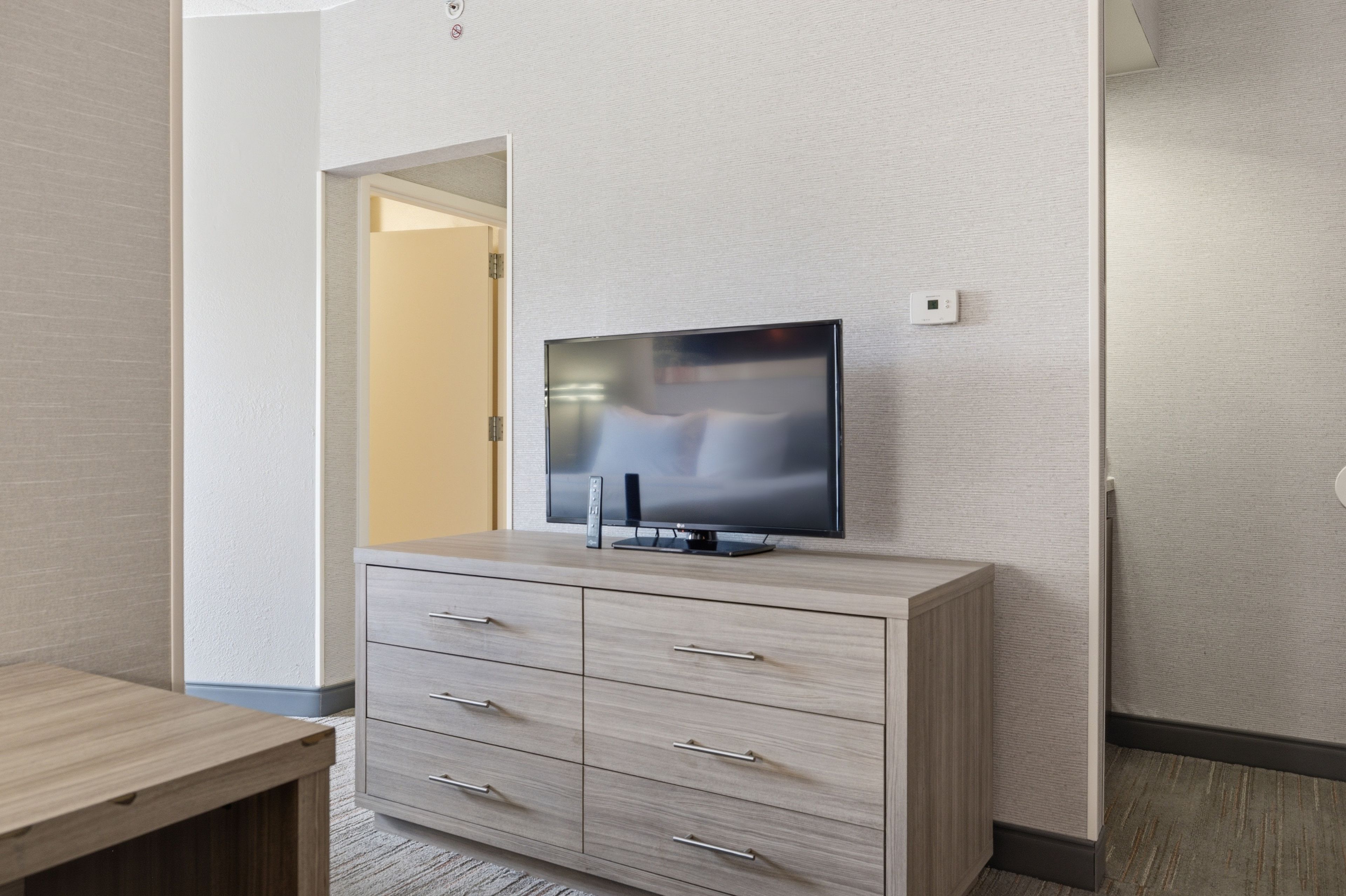 Una habitación moderna con televisor de pantalla plana sobre un mueble de madera, una puerta que da a otra habitación y un termostato en la pared.