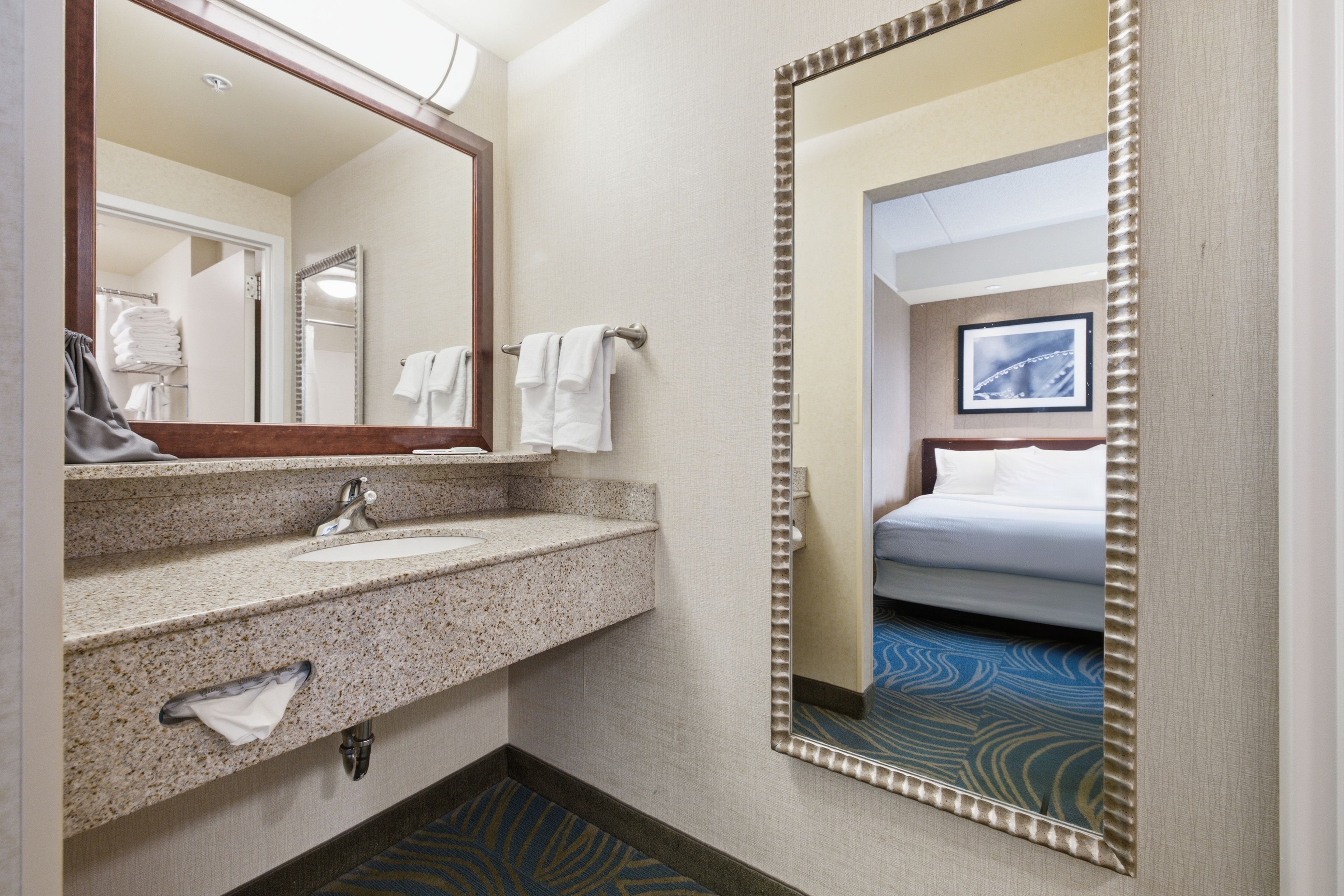 A hotel bathroom with a granite sink, a mirror, and a bed visible through an adjacent door.