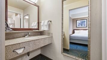 Un baño de hotel con un lavamanos de granito, un espejo y una cama visible a través de una puerta contigua.