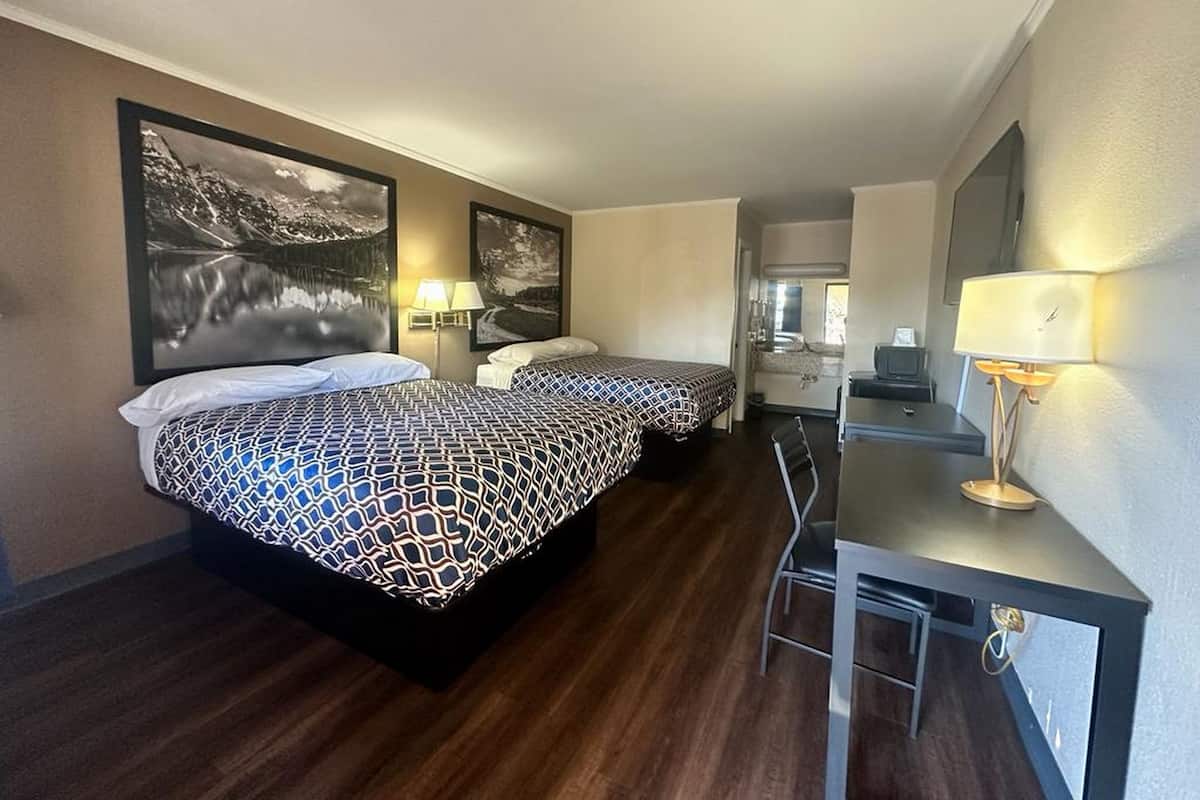 Une chambre d’hôtel avec deux lits, un bureau et une chaise. Il y a deux lampes fixées au mur et un grand tableau encadré accroché au mur.