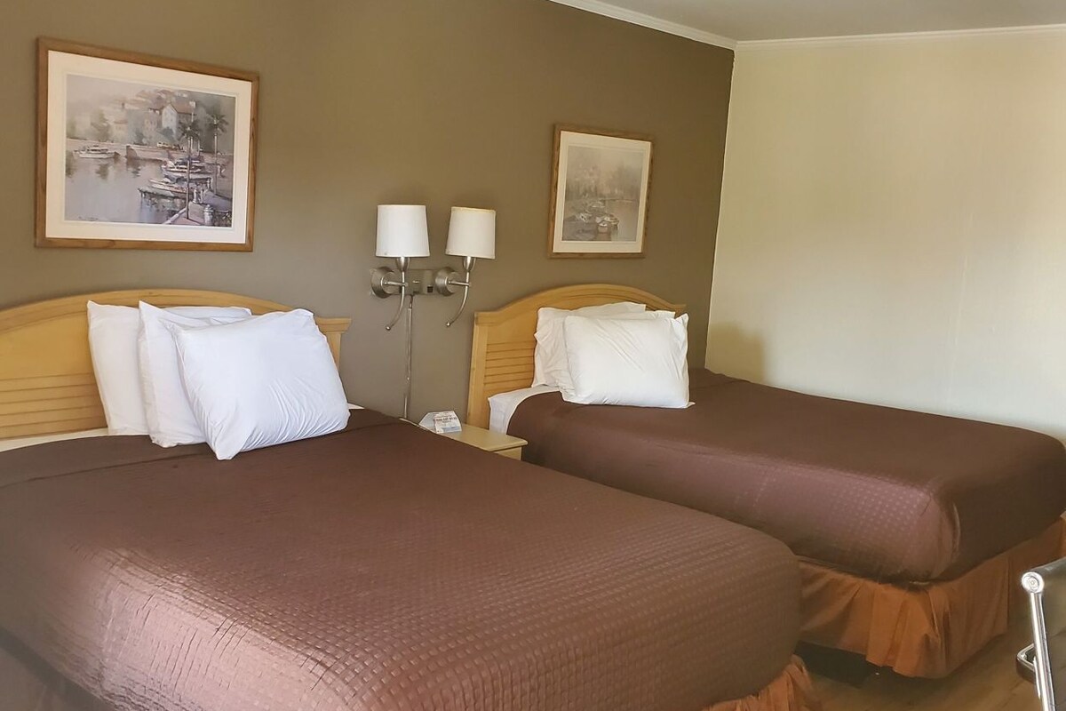 Quarto de hotel com duas camas, piso de madeira e duas luminárias na parede.