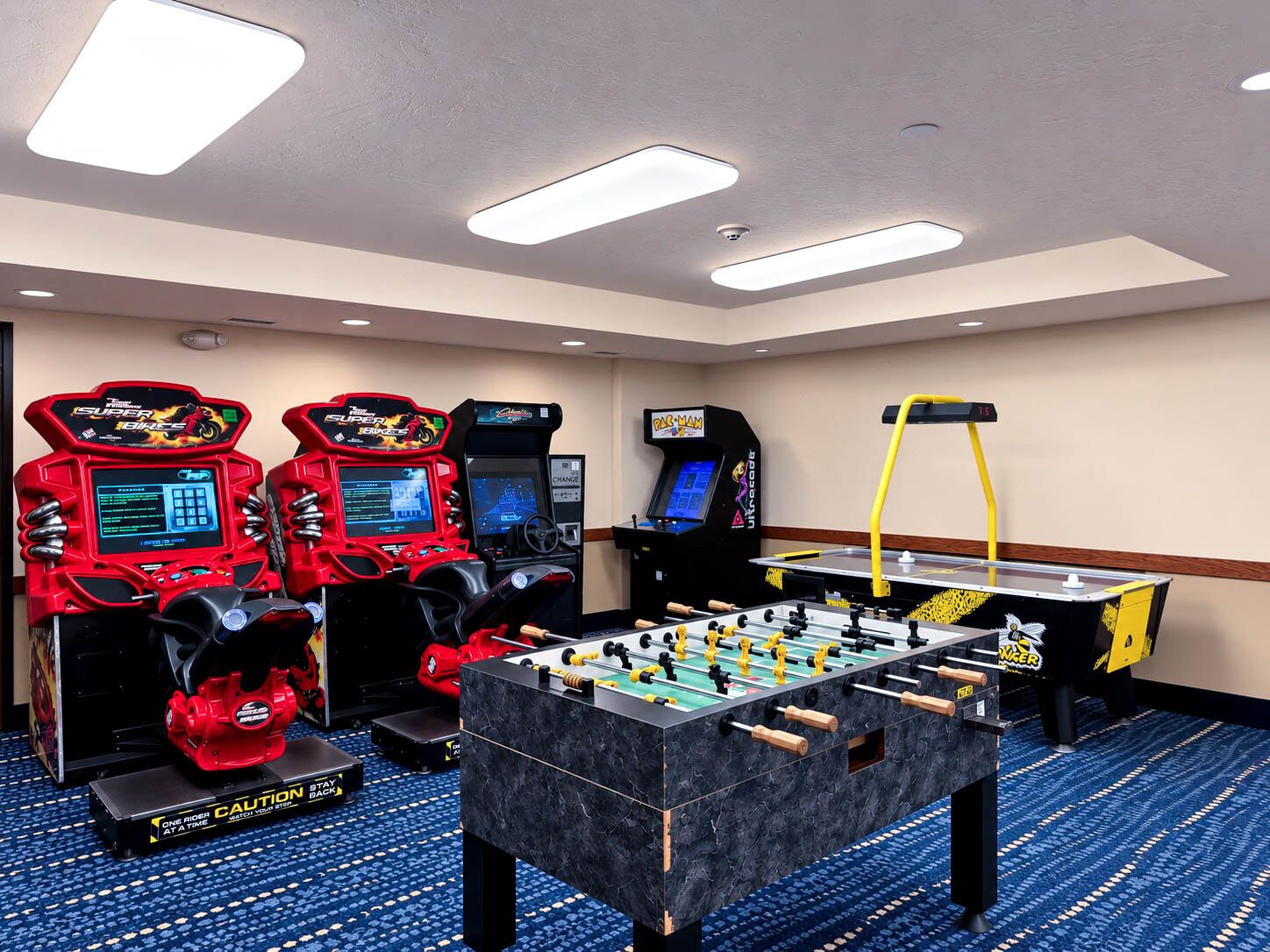 Una habitación con cuatro máquinas de arcade, una mesa de fútbol de mesa y una máquina de ejercicios amarilla.