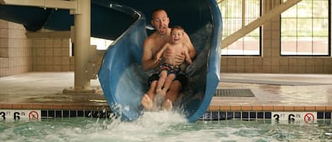 A man and a child sliding down a water slide in an indoor pool area.