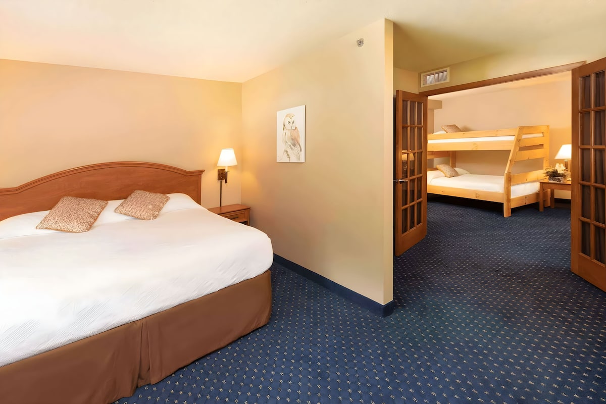 Ein Hotelzimmer mit einem großen Bett, einem Nachttisch mit Lampe, einem Wandbild und einem Stockbett im angrenzenden Zimmer.