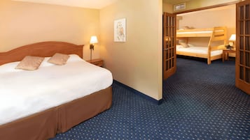 A hotel room with a large bed, a bedside table with a lamp, a wall-mounted artwork, and a bunk bed in the adjacent room.