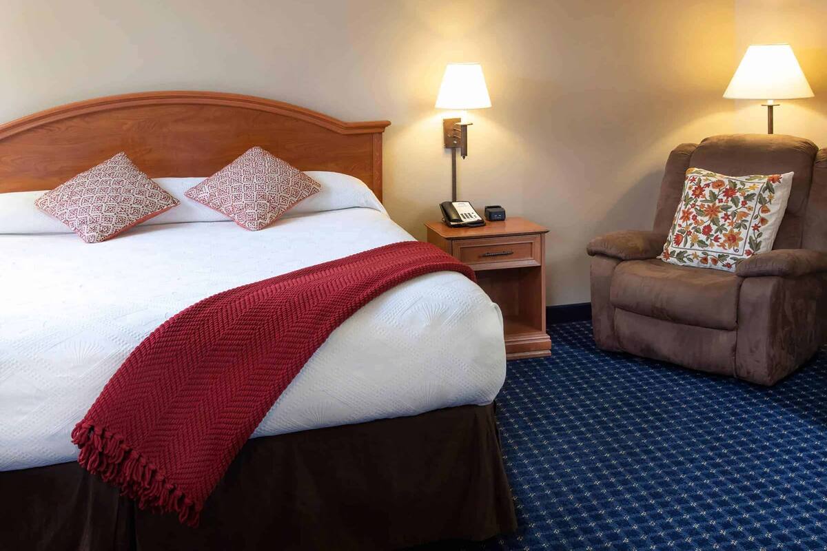 Ein Hotelzimmer mit einem Bett, zwei Kissen, einer roten Tagesdecke, einem Nachttisch mit Lampe, einem Telefon und einem Sessel mit Blumenkissen.