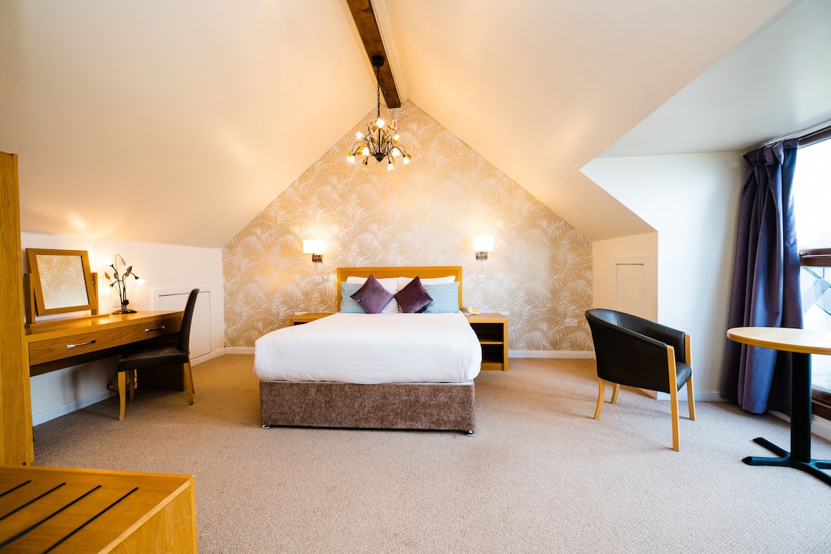 A bedroom with a sloped ceiling, a bed with a wooden headboard, a desk with a chair, a small table, and a chandelier.