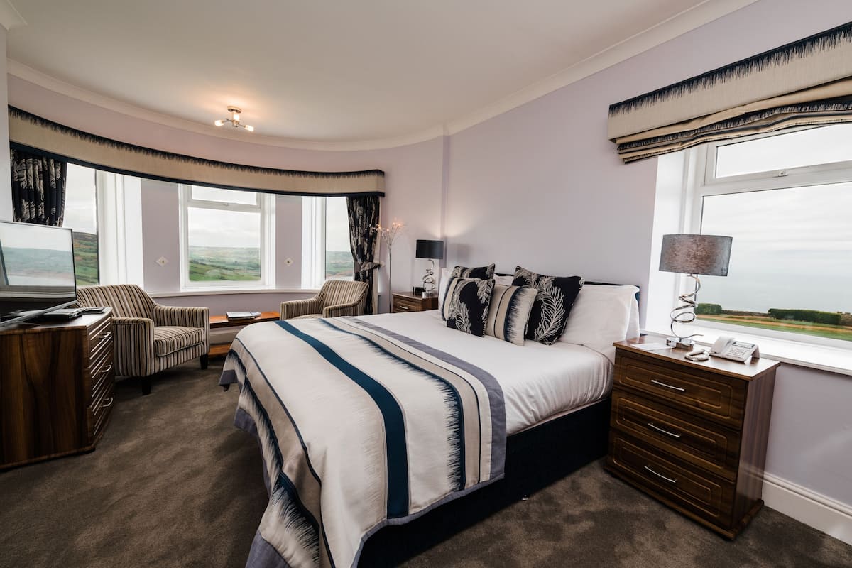A bedroom with a large bed, a desk, a chair, and a nightstand with a lamp. There is a window with a view of the landscape.