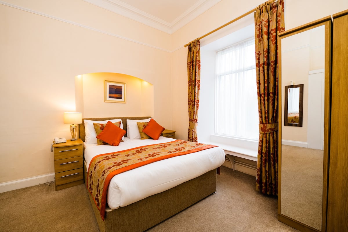 A neatly arranged bedroom with a large bed, a bedside table with a lamp, a mirror, and a window with curtains.