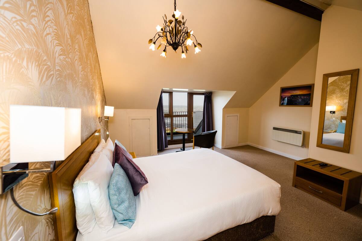 A bedroom with a large bed, a chandelier, a view of the outside, a small table, and a chair.