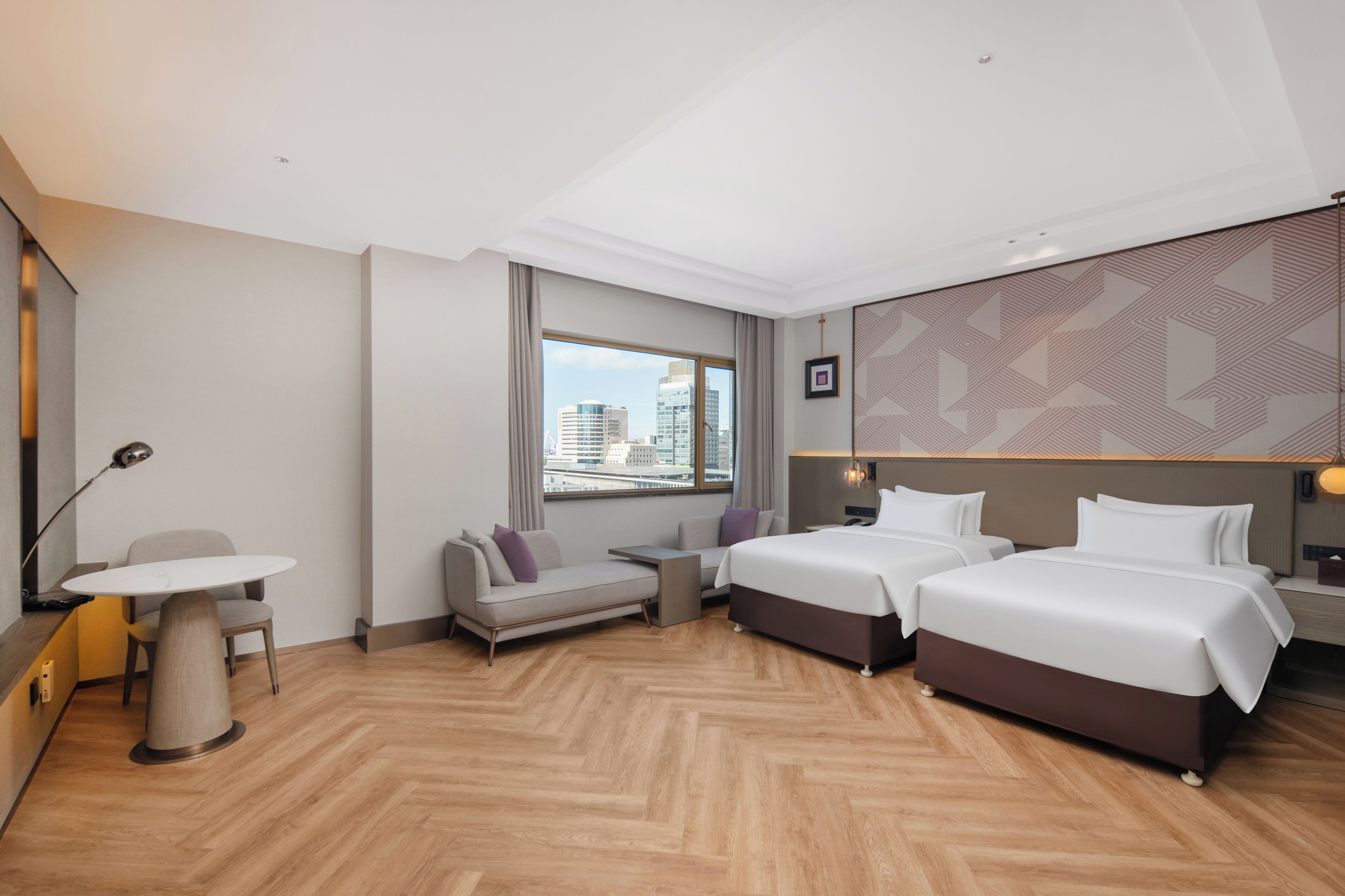 Een hotelkamer met twee bedden, een bureau, een stoel, een bank en een groot raam met uitzicht op de stad.