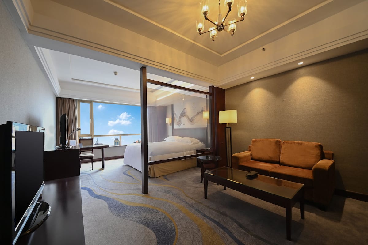 Habitación de hotel con cama, sofá, escritorio, televisor y un ventanal con vistas al cielo.