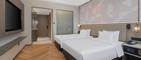 A modern hotel room with two beds, a flat-screen TV, a bedside table with a lamp, and a large mirror.