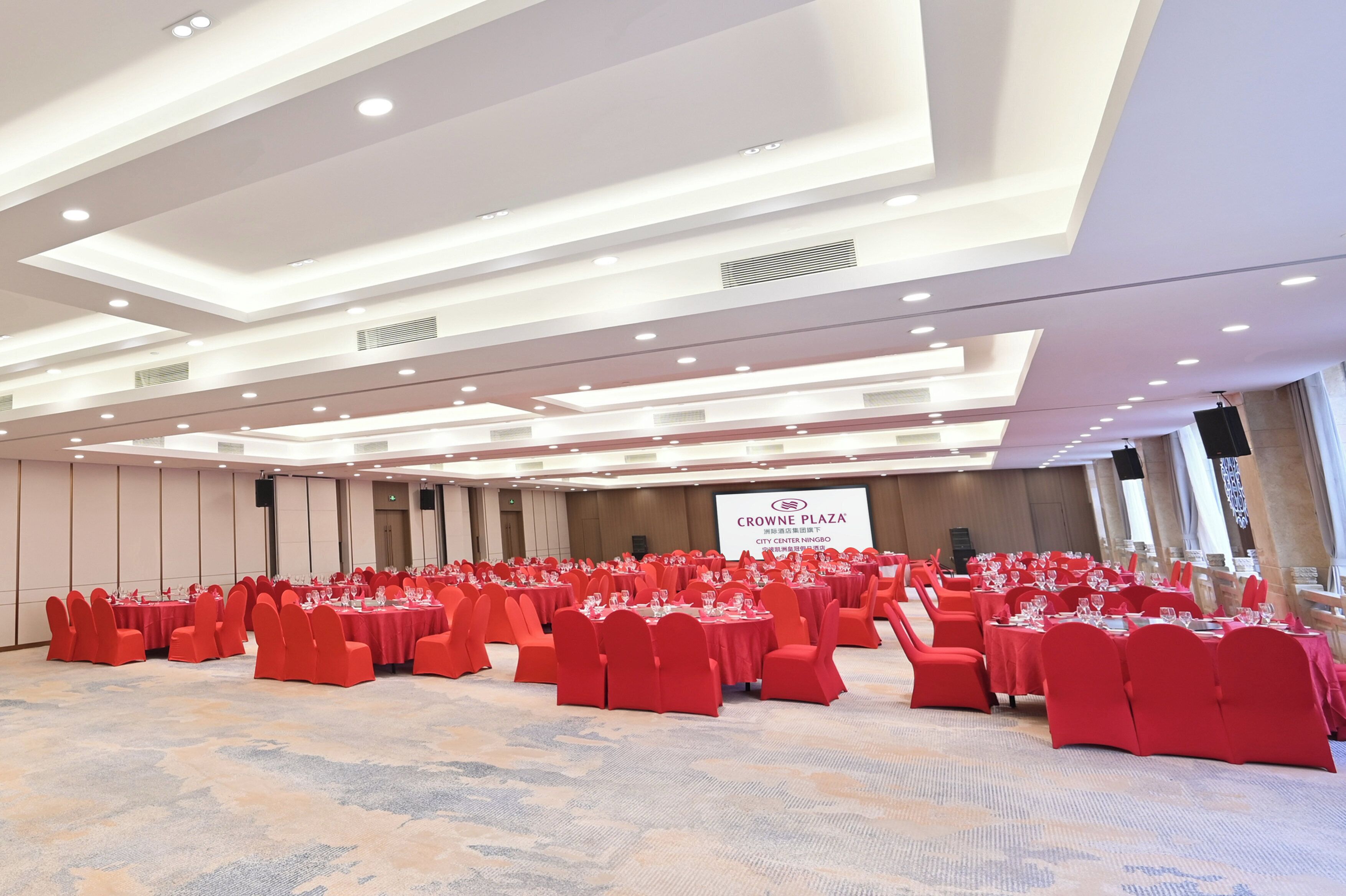 A conference room with multiple tables and chairs, a large screen displaying text, and a carpeted floor.