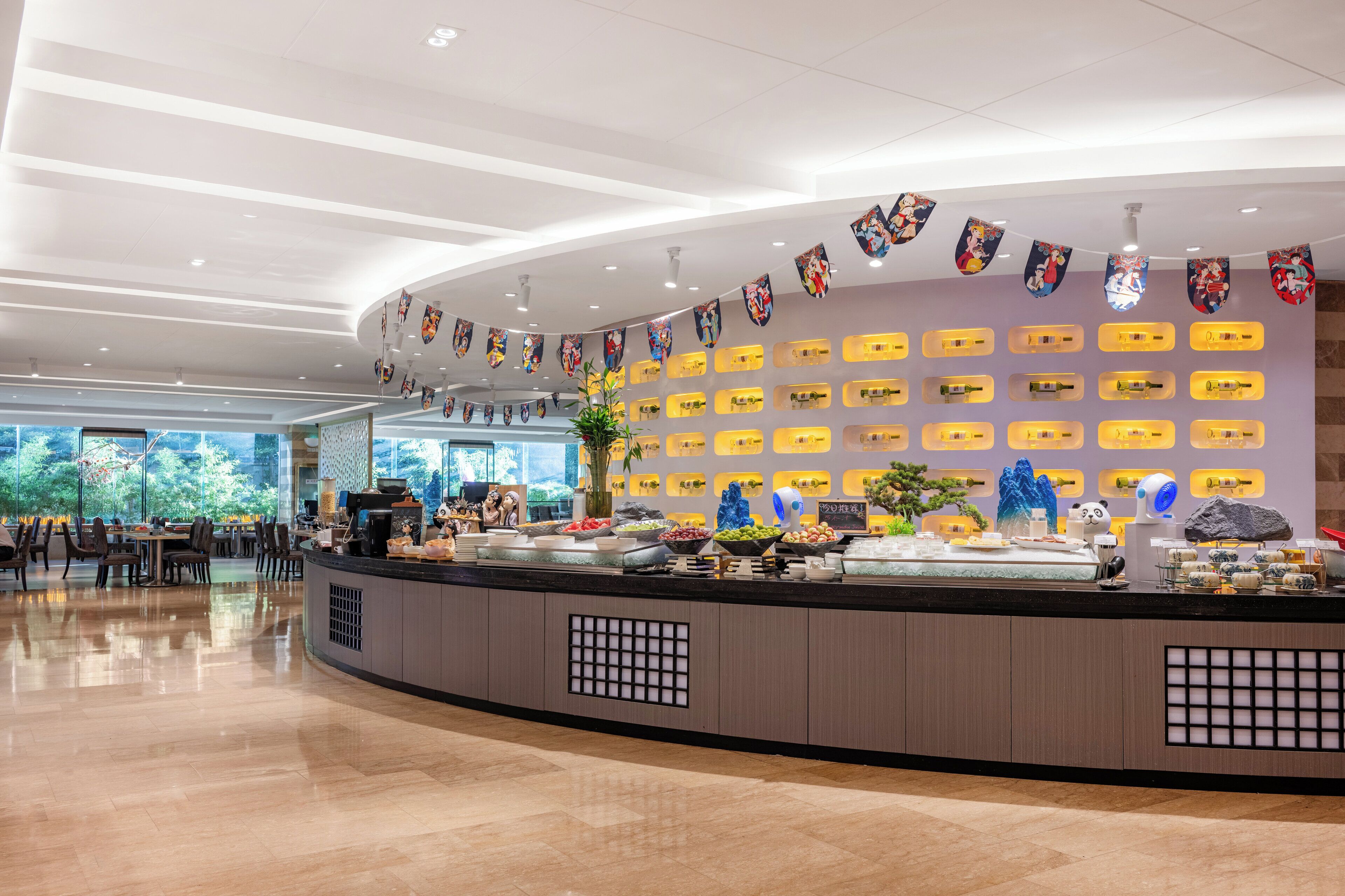 A modern hotel lobby with a curved reception desk, a variety of food displays, and a wall decorated with colorful artwork.